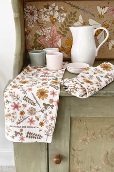 a table topped with a white pitcher and two cups on top of a wooden cabinet