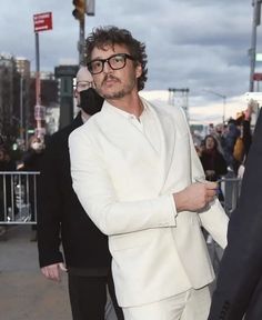 a man in a white suit and glasses walking down the street with other people behind him