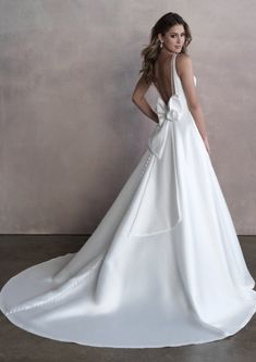 a woman in a white wedding dress with a bow on her back, standing against a wall