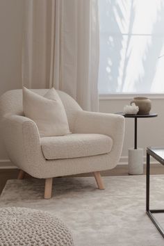 a white chair sitting in front of a window next to a table with a vase on it