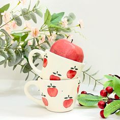 two mugs with apples painted on them are next to some flowers and leaves in the background