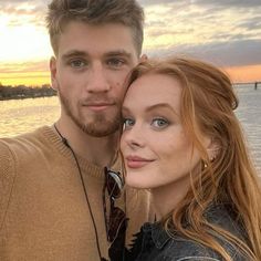 a man and woman standing next to each other near the water at sunset or sunrise