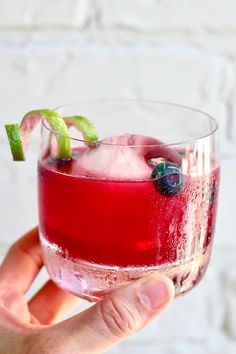 a hand holding a wine glass filled with ice and blueberries on the rim, in front of a white brick wall