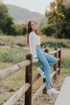 a young woman sitting on top of a wooden fence next to a lush green field