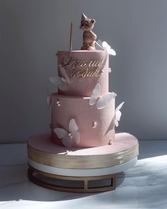 a three tiered pink cake with white butterflies and a cat figurine on top