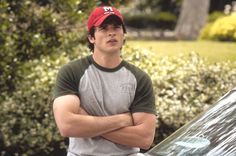 a man with his arms crossed standing in front of a car wearing a red hat