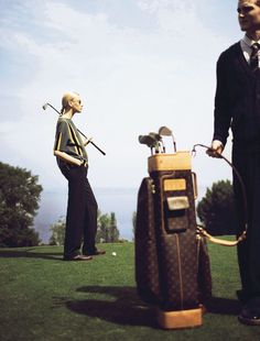 a man standing next to a woman holding golf clubs and a bag on top of a green field