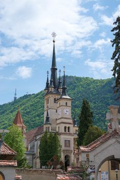 an old church with steeples and clocks on the front is surrounded by trees, bushes, and mountains