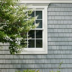 a cat sitting on the window sill of a gray house next to bushes and trees