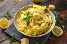 a bowl of pasta with lemons and basil on a wooden table next to bread