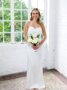 a woman standing in front of a window wearing a white dress and holding a bouquet