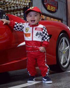 a little boy standing in front of a red sports car wearing a checkered outfit