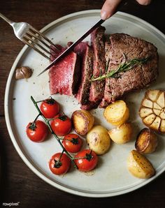 a person cutting up some meat on top of a plate with potatoes and cherry tomatoes