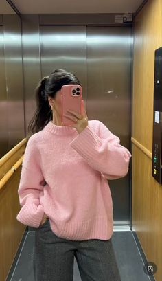 a woman taking a selfie in an elevator wearing a pink sweater and grey pants