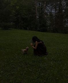 a woman kneeling down in the grass with a small dog next to her and trees in the background