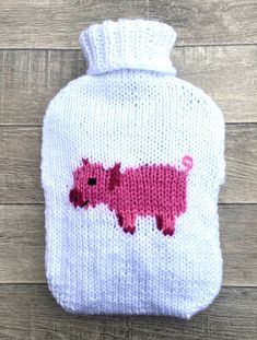 a white knitted hot water bottle with pink pigs on the front and bottom, sitting on a wooden surface