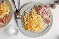 two plates with pasta, ham and cheese on them next to a fork and knife