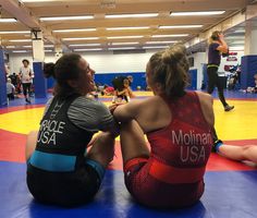 two women sitting on the ground talking to each other in an indoor wrestling ring while others watch