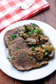 two steaks with mushrooms on a white plate next to a red and white checkered napkin