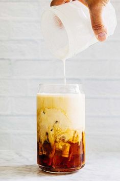 someone pours milk into a glass filled with ice cream and brown liquid on a white surface