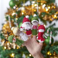 a hand holding three knitted christmas ornaments in front of a christmas tree