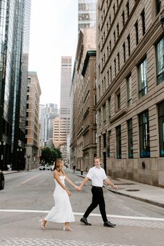 a man and woman holding hands crossing the street in front of tall buildings on a city street