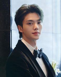 a young man in a tuxedo and bow tie looking at the camera while standing next to a wall