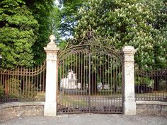 an iron gate in the middle of a stone wall with a statue on it and trees behind it