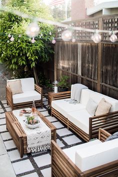 an outdoor patio with white cushions and wooden furniture on the ground, in front of a fence