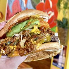 a hand holding a sandwich with meat, cheese and veggies on it in front of a colorful wall