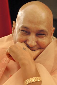 a balding man wearing a pink shirt and gold bracelet smiles at the camera with his chin resting on his hand