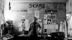 black and white photograph of man standing in front of wall with posters on it,