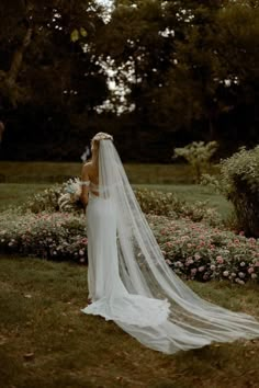 a woman in a wedding dress and veil standing on the grass with flowers behind her