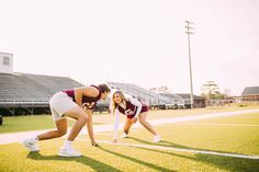 two young women in cheerleader outfits on a football field, one kneeling down to pick up the ball