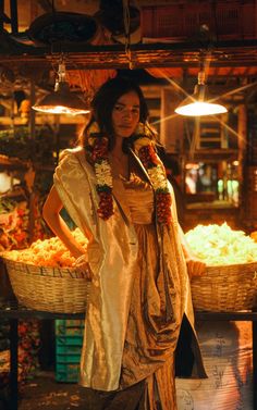 a woman standing in front of a basket full of fruit at an outdoor market with lights hanging from the ceiling