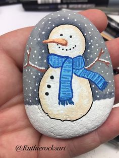 a hand holding a painted rock with a snowman wearing a blue hat and scarf