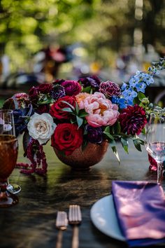 an arrangement of flowers in a basket on a table with wine glasses and napkins