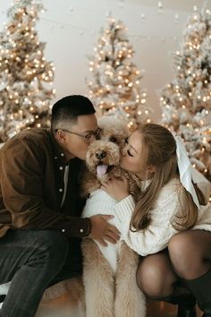 a man and woman kissing while holding a dog in front of christmas trees with lights