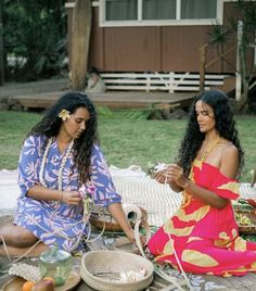 two women sitting on the ground in front of a picnic table with fruit and flowers