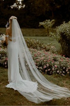 a woman in a wedding dress and veil walking through the grass with flowers behind her