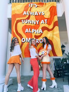 three women in orange outfits standing next to an orange and yellow sign that says it's always sunny at alpha omro