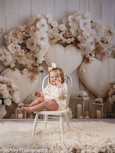 a baby sitting on a chair in front of a backdrop with white flowers and hearts