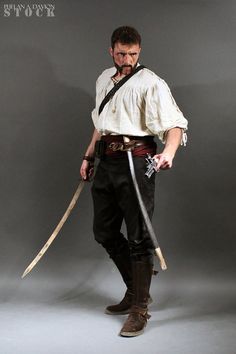 a man dressed in pirate garb holding two swords and looking at the camera while standing against a gray background