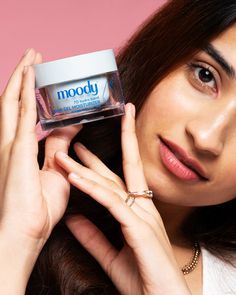 a woman holding up a jar of moody moisturizing cream on her face