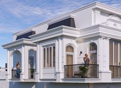 two people standing on the balcony of a white house with black roof and balconies