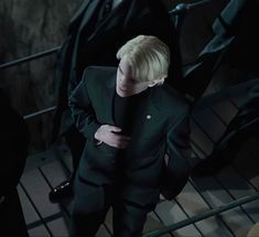 a blonde haired man in a black suit looking at his cell phone while standing on stairs