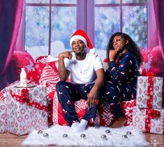 a man and woman sitting on presents in front of a christmas tree with santa's hat