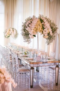 a long table with clear chairs and white flowers on the top is surrounded by tall centerpieces