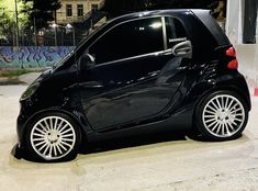 a small black car parked in front of a wall with graffiti on it's side