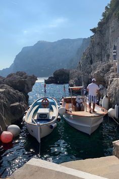 two small boats tied to the shore with people standing on one boat in the water